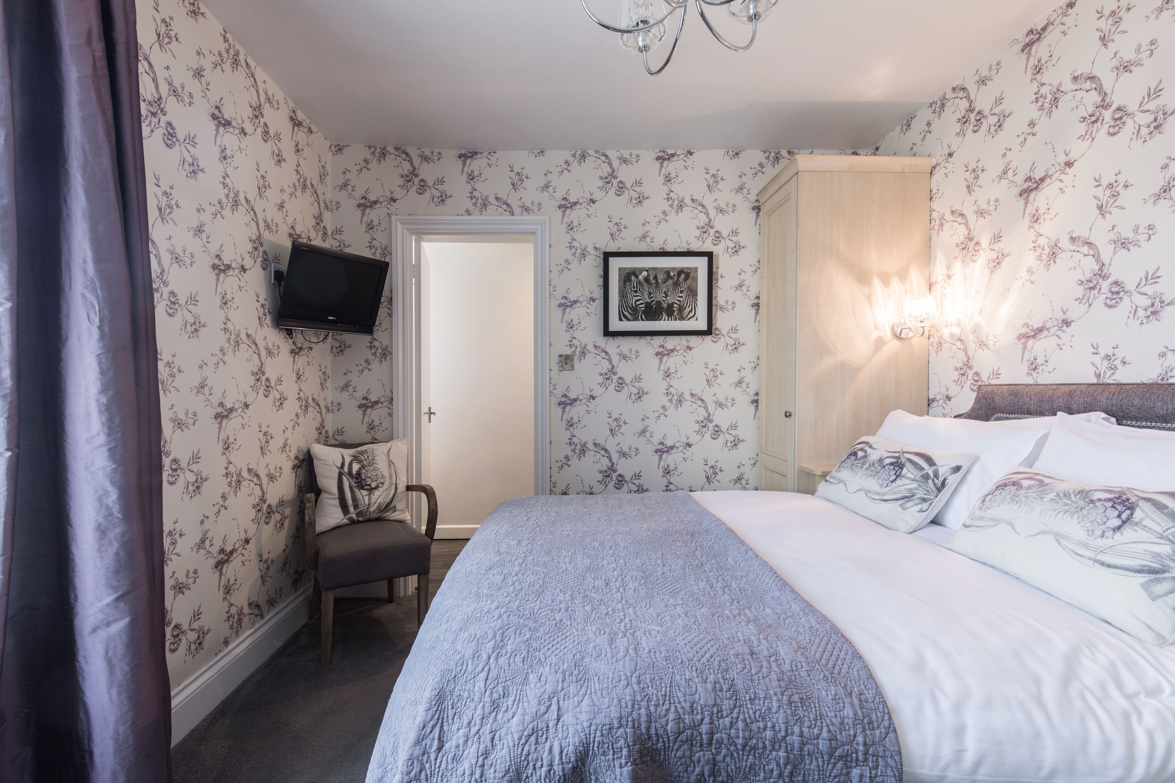 Een slaapkamer met bloemenbehang, een bed met opvallende kussens, een stoel, een flatscreen televisie en een deur.