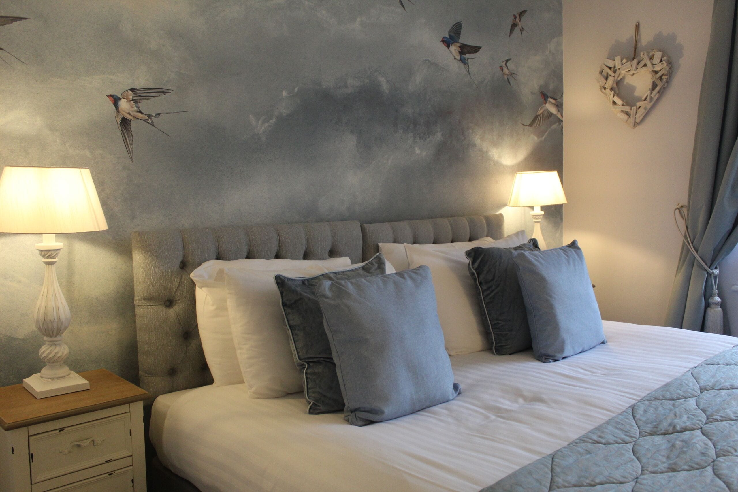 Ein Schlafzimmer mit einem großen Bett, zwei Nachttischlampen, einem Nachttisch und einem Wandbild mit Vögeln.