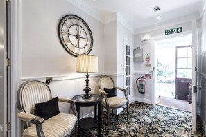 Un pasillo con un gran reloj de pared, dos sillones tapizados, una mesita con una lámpara, un letrero de salida de incendios y un extintor.