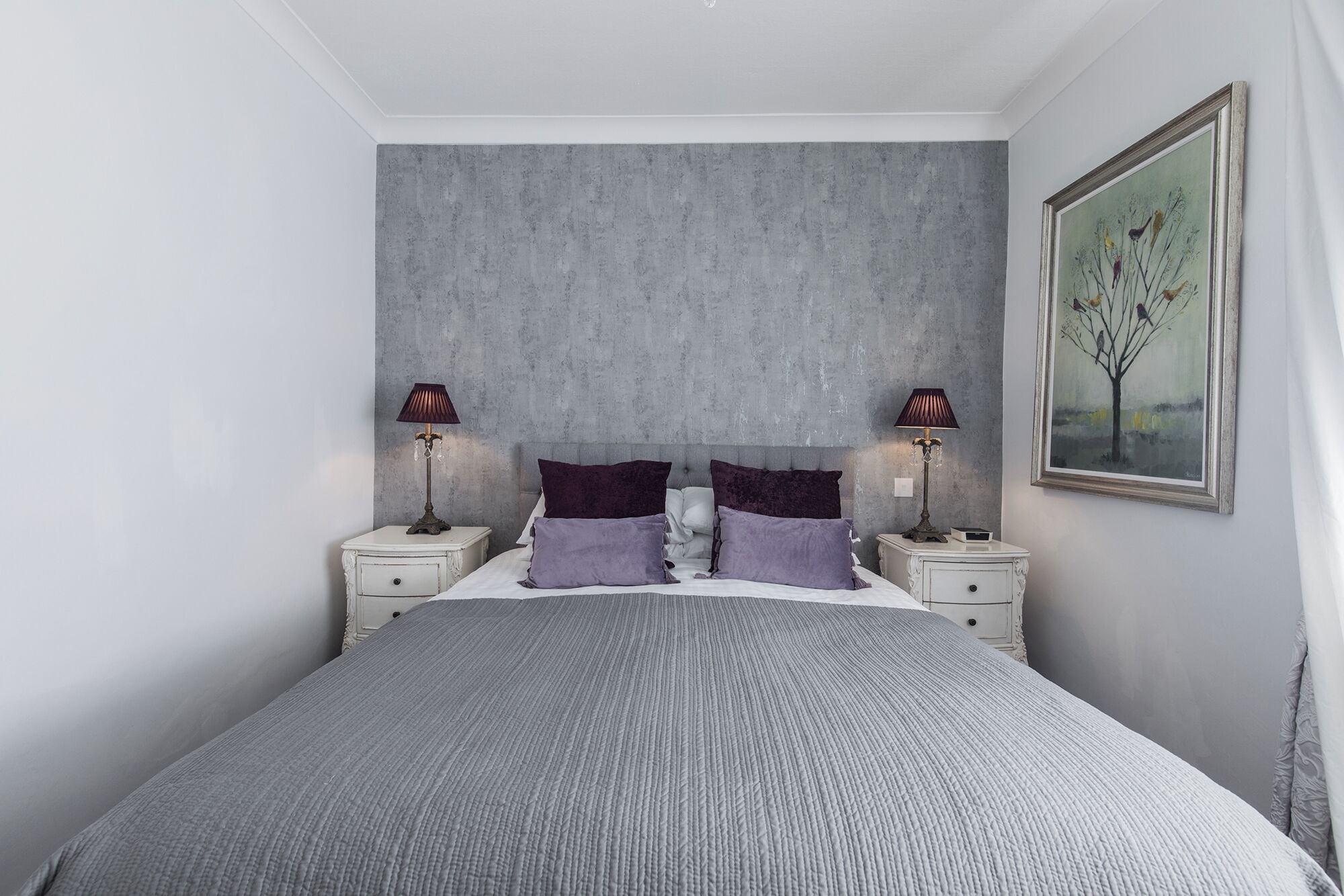 Een slaapkamer met een groot bed, twee nachtkastjes, een grijs geribbeld hoofdbord en een lijst met een schilderij aan de muur.