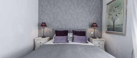 A bedroom with a large bed, two nightstands, a grey textured headboard, and a framed painting on the wall.