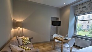 Una sala de estar moderna con un sofá, una mesa de comedor con sillas, un televisor montado en la pared y una ventana con vistas a edificios.