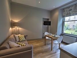 Una sala de estar moderna con un sofá, una mesa de comedor con sillas, un televisor montado en la pared y una ventana con vistas a edificios.