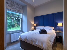 A bedroom with a large window, a blue headboard, two bedside tables with lamps, and a neatly made bed with rolled towels.