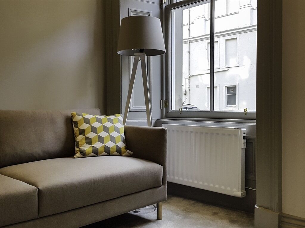 Une pièce comprenant un canapé, un coussin à motifs, une lampe sur pied et une fenêtre donnant sur les bâtiments environnants.