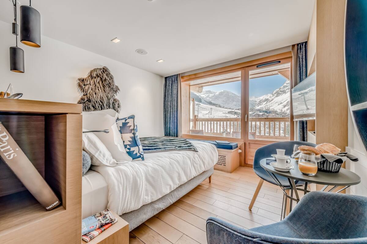 Una habitación de hotel moderna con cama, una mesita con desayuno, una silla y un ventanal con vistas a la montaña.