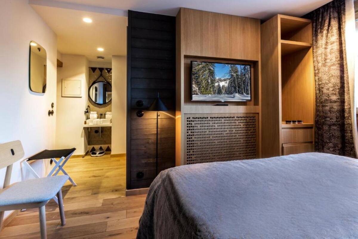 Un dormitorio moderno con cama, televisor montado en un panel de madera, un pequeño rincón de descanso y un baño con espejo y lavamanos.