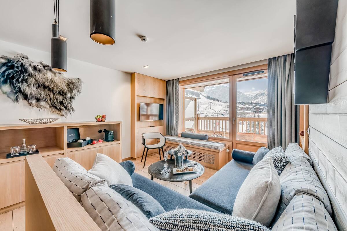 Una sala de estar moderna con un sofá, una mesita redonda y vistas a un paisaje montañoso nevado a través de grandes ventanales.