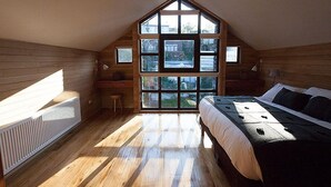 Una camera da letto con soffitto spiovente, pareti in legno, un'ampia finestra, un letto, una scrivania e un termosifone.