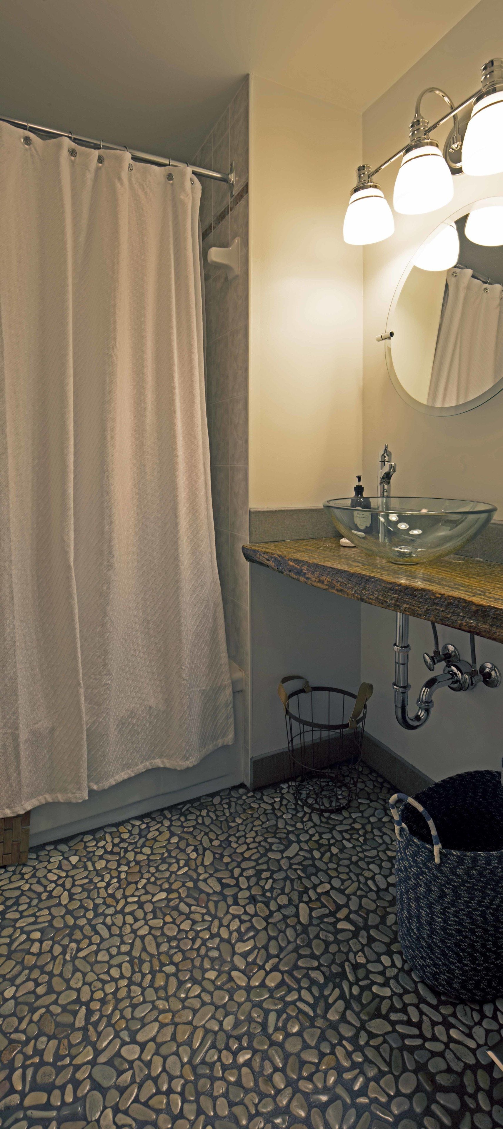 A bathroom with a glass sink, a shower curtain, a mirror, and a pebbled floor.