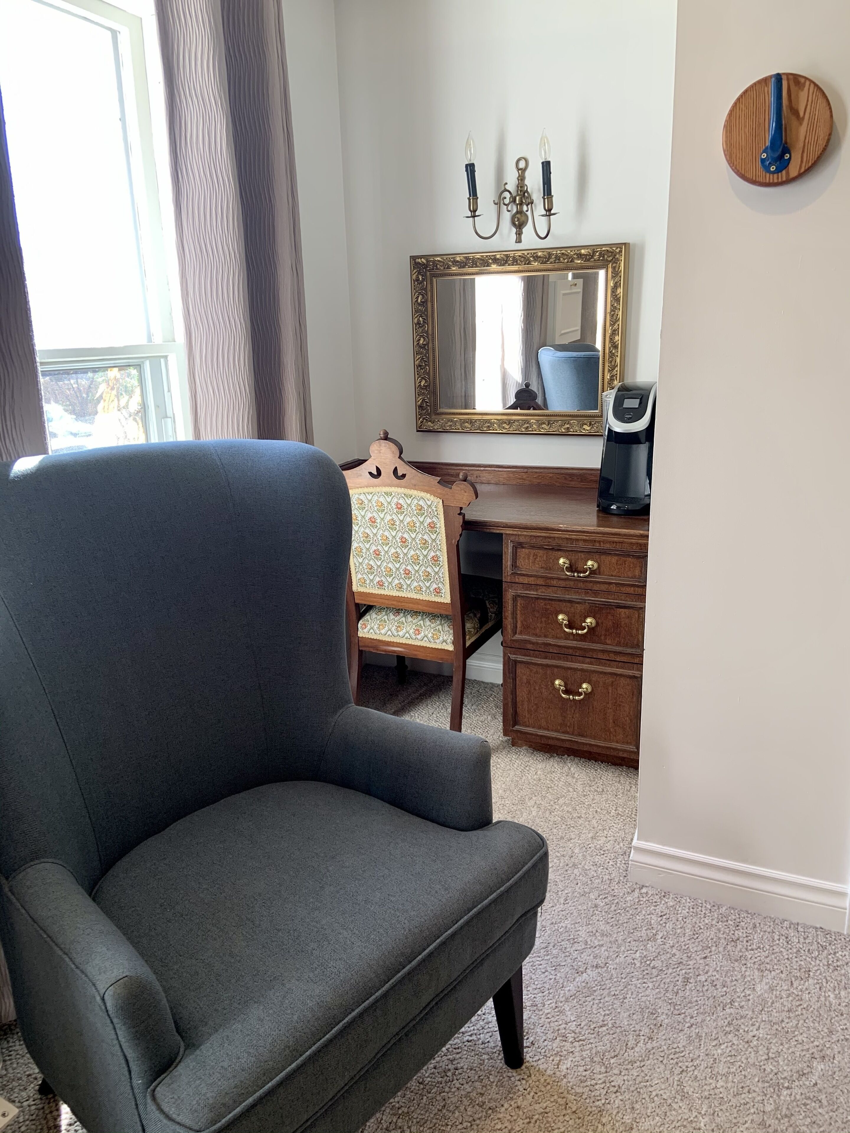 A room with a grey armchair, a wooden desk with a coffee maker, a mirror, and a wall-mounted clock.