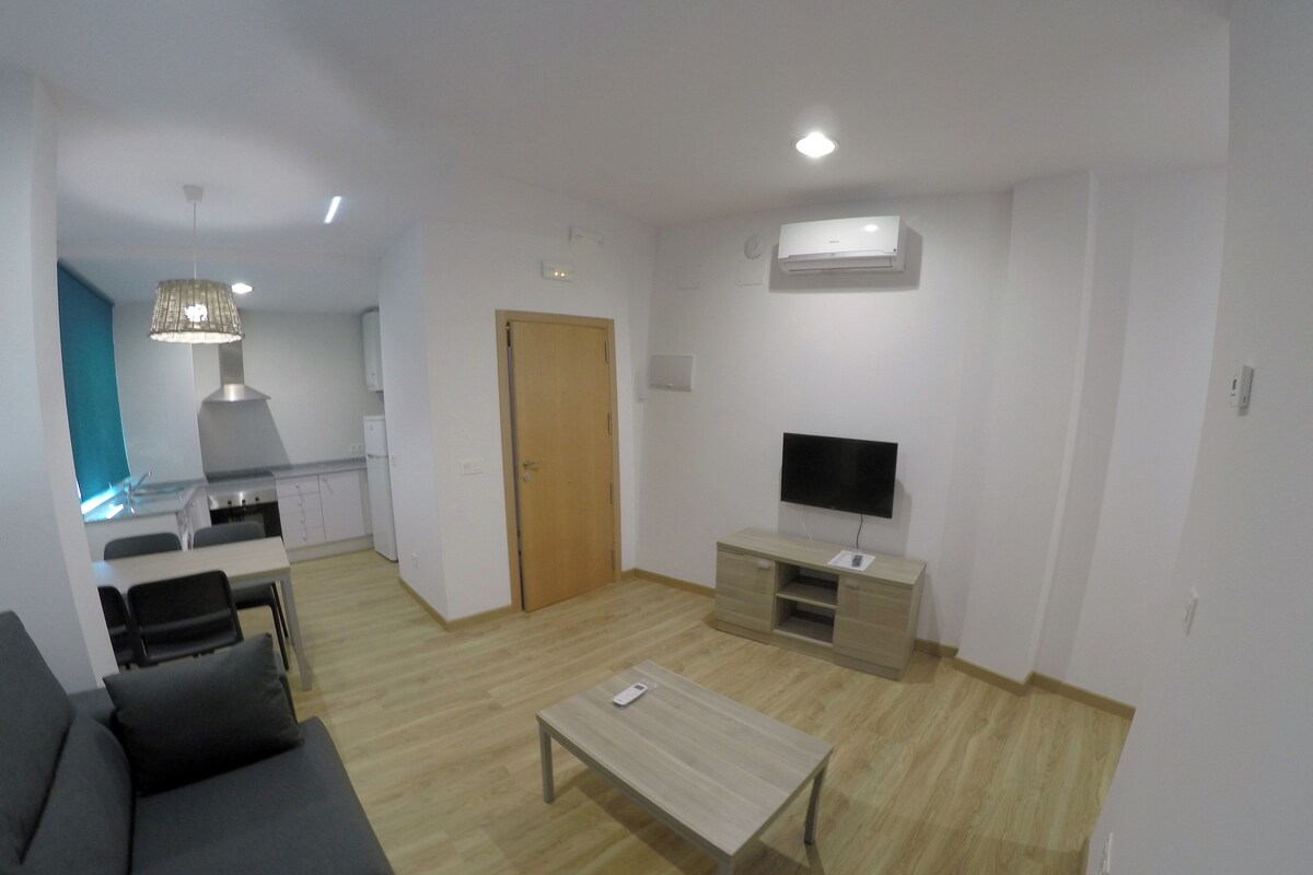 Una sala de estar moderna con un sofá, una mesa de centro, un televisor y una cocina al fondo.