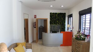 A hotel lobby with a reception desk, a sofa, a cabinet, and a wall with a green leafy pattern. - Hotel Sagunto (Sagunto)