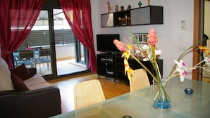 A living room with a sofa, a glass table, a vase with flowers, a television, and a sliding glass door with curtains. - Apartamento Puerto Rico 39 (Tossa de Mar)