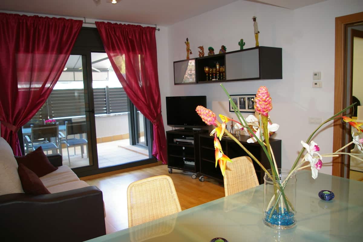 Uma sala de estar com sofá, mesa de vidro, vaso com flores, televisão e uma porta de correr com cortinas.