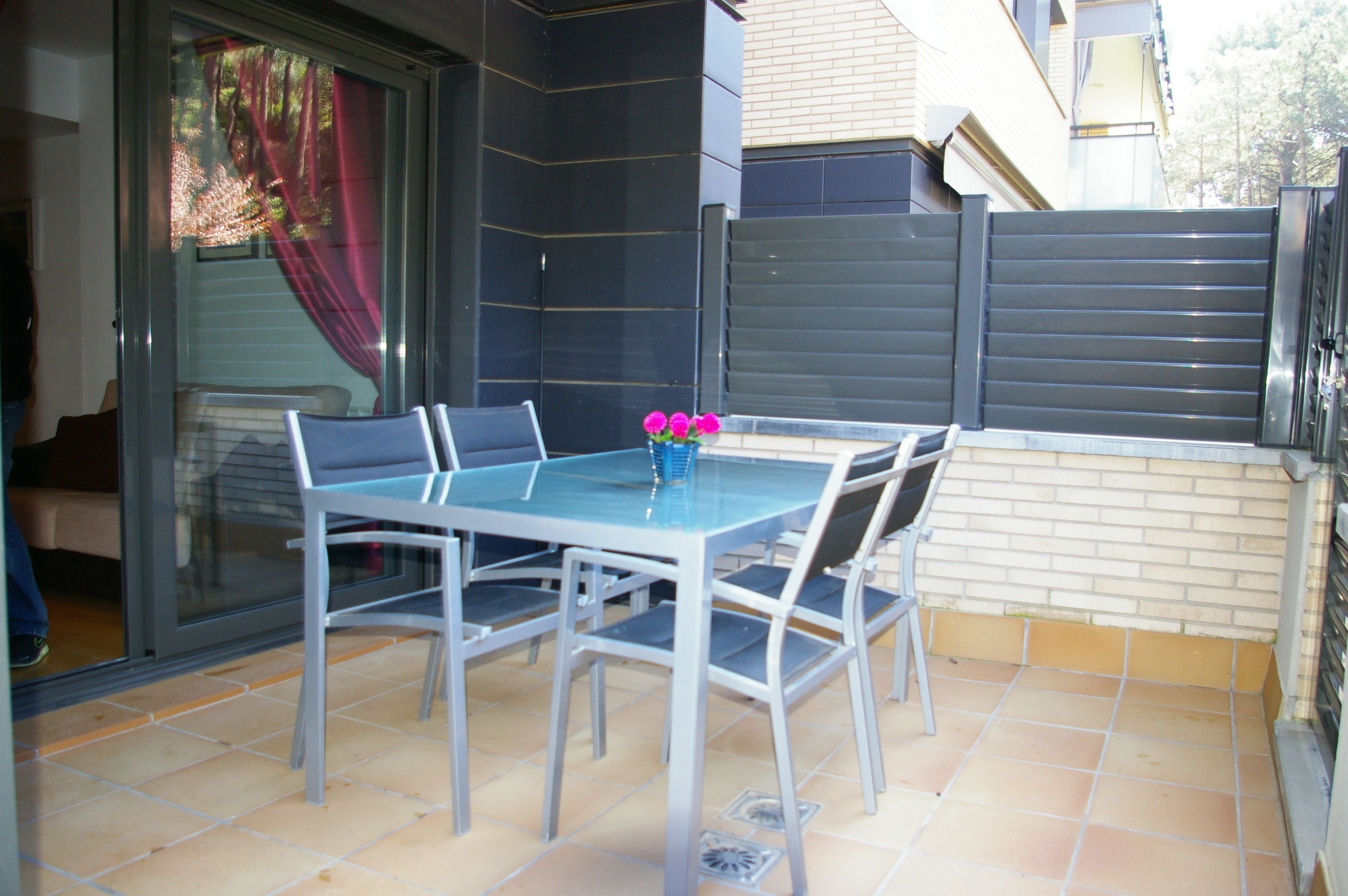 Un patio con una mesa y sillas de vidrio, un jarrón pequeño con flores y un ventanal corredero.