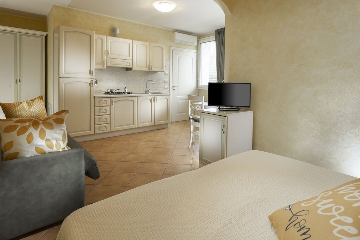 A compact hotel room with a bed, a television, a kitchenette, and a seating area.