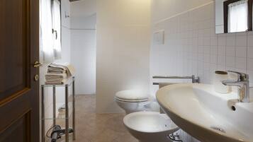 Un bagno moderno con WC, bidet e lavandino. È presente un porta asciugamani con asciugamani piegati e uno scaffale in vetro con un asciugacapelli.