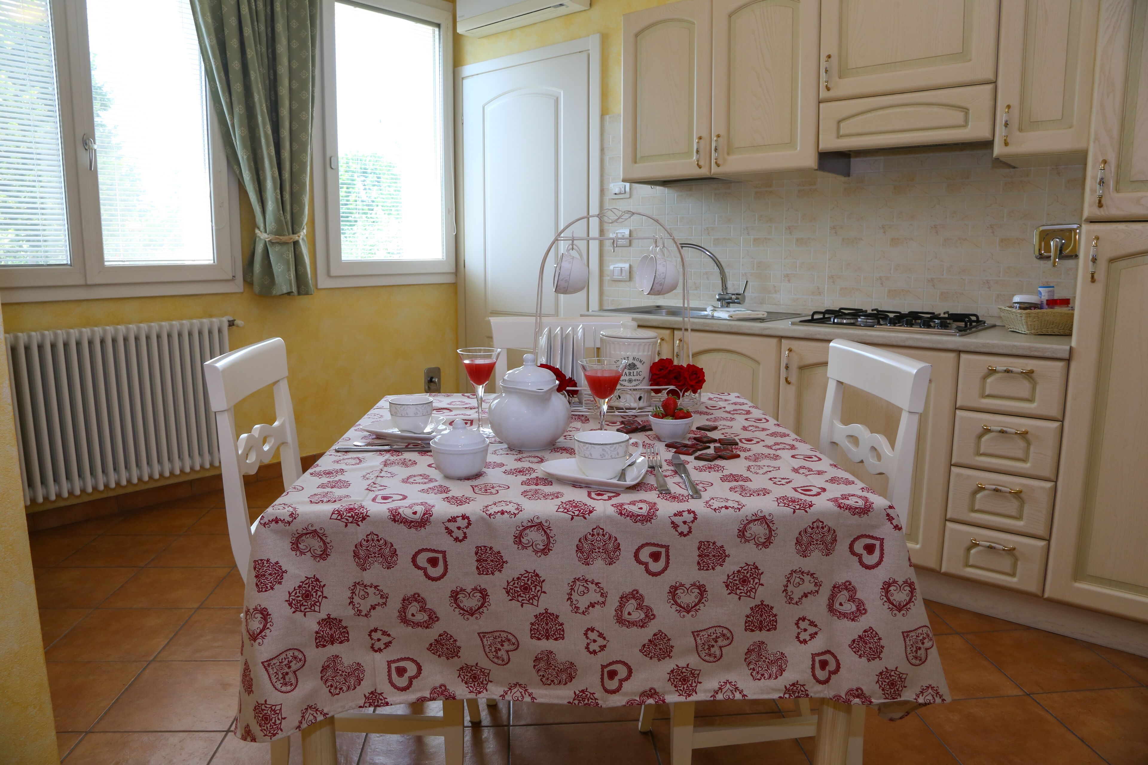 Una cocina con una mesa de comedor preparada para dos personas, armarios blancos, una ventana con cortinas y un salpicadero de azulejos.