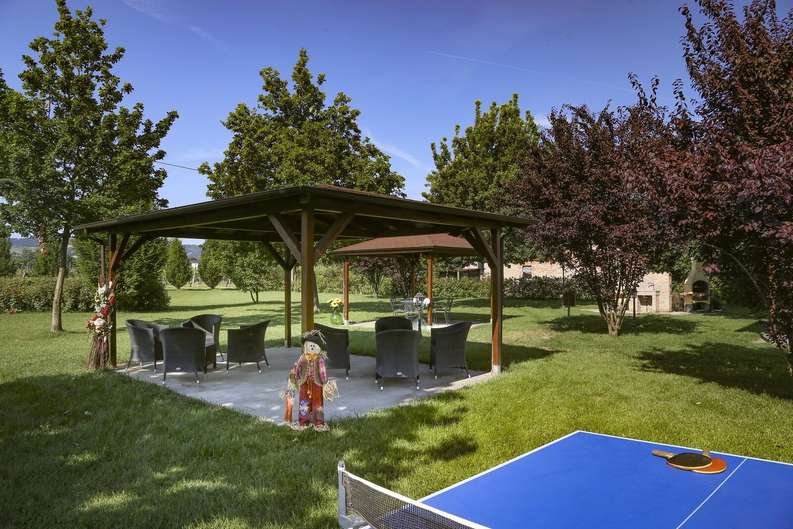A covered outdoor seating area with chairs and a table, surrounded by grass and trees.
