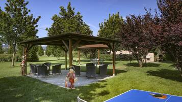 A covered outdoor seating area with chairs and a table, surrounded by grass and trees.