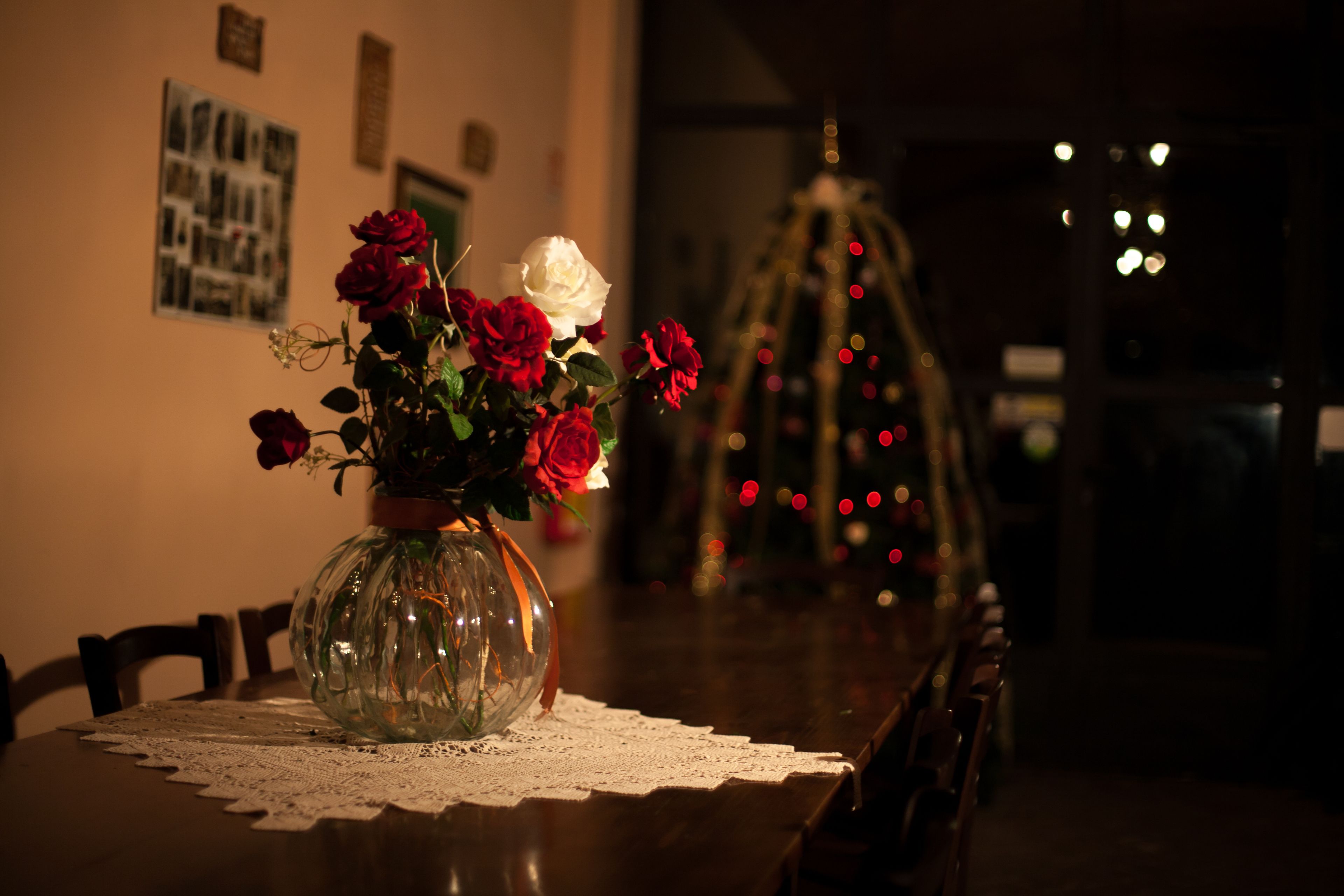 A dining table with a lace tablecloth, a vase with red and white roses, and a Christmas tree in the background.