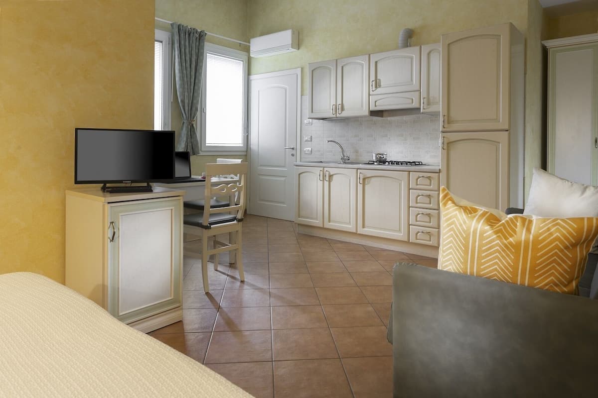 A compact hotel room with a bed, a desk with a TV, a chair, a sofa, and a kitchenette with cabinets and a stove.