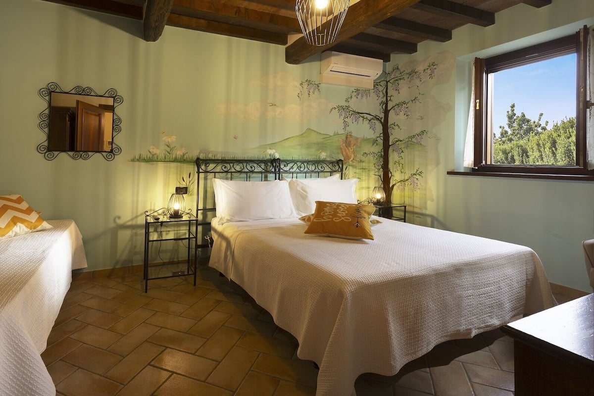 A bedroom with a bed, nightstands, a mirror, and a window with a view of trees.