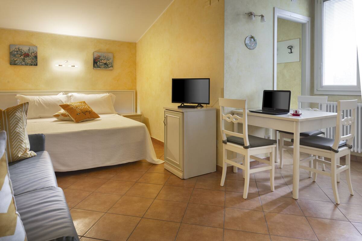 A bedroom with a bed, a desk with a laptop, a chair, a small cabinet, and a window with blinds.