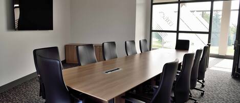 Uma sala de conferência com uma mesa comprida, cadeiras pretas, uma TV de tela plana e grandes janelas com vista para um edifício.