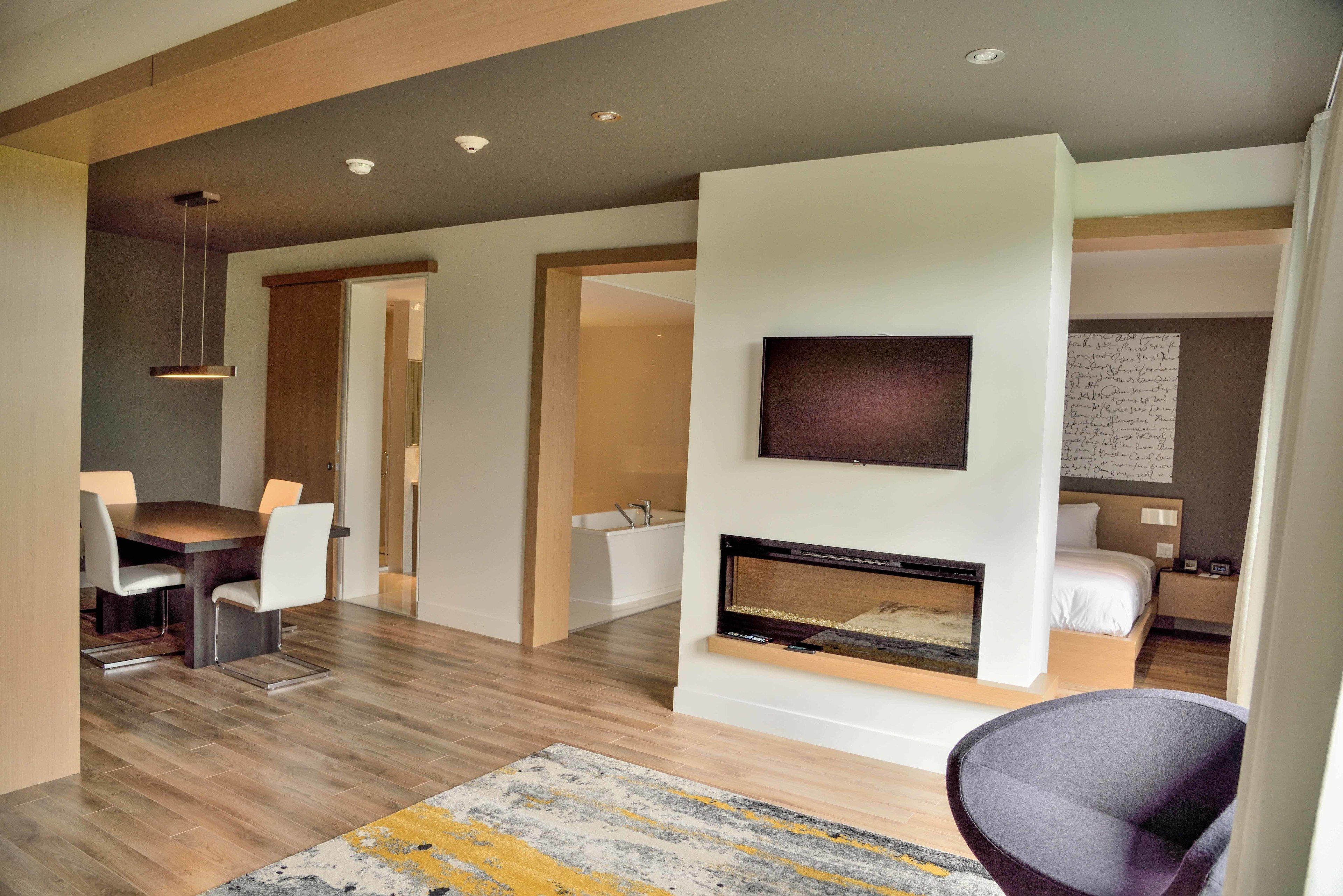 A modern hotel room with a flat-screen TV mounted on the wall, a fireplace, a dining area with a table and chairs, and a bed with a nightstand.