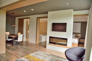 Quarto de hotel moderno com televisão de tela plana na parede, lareira, área de jantar com mesa e cadeiras, e uma cama com mesa de cabeceira.