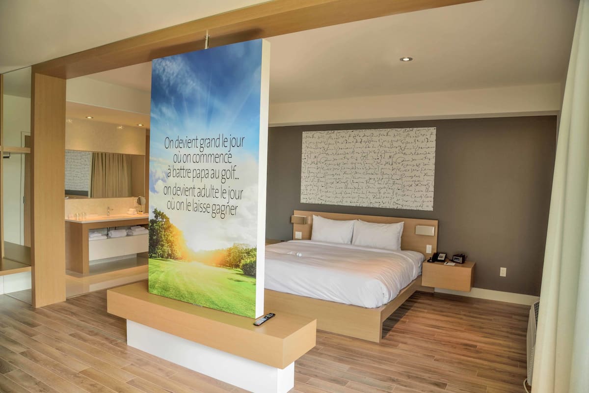 Een moderne hotelkamer met een groot bed, een nachtkastje met lamp, een aan de muur hangend kunstwerk, en een badkamer met wastafel en handdoekrek.