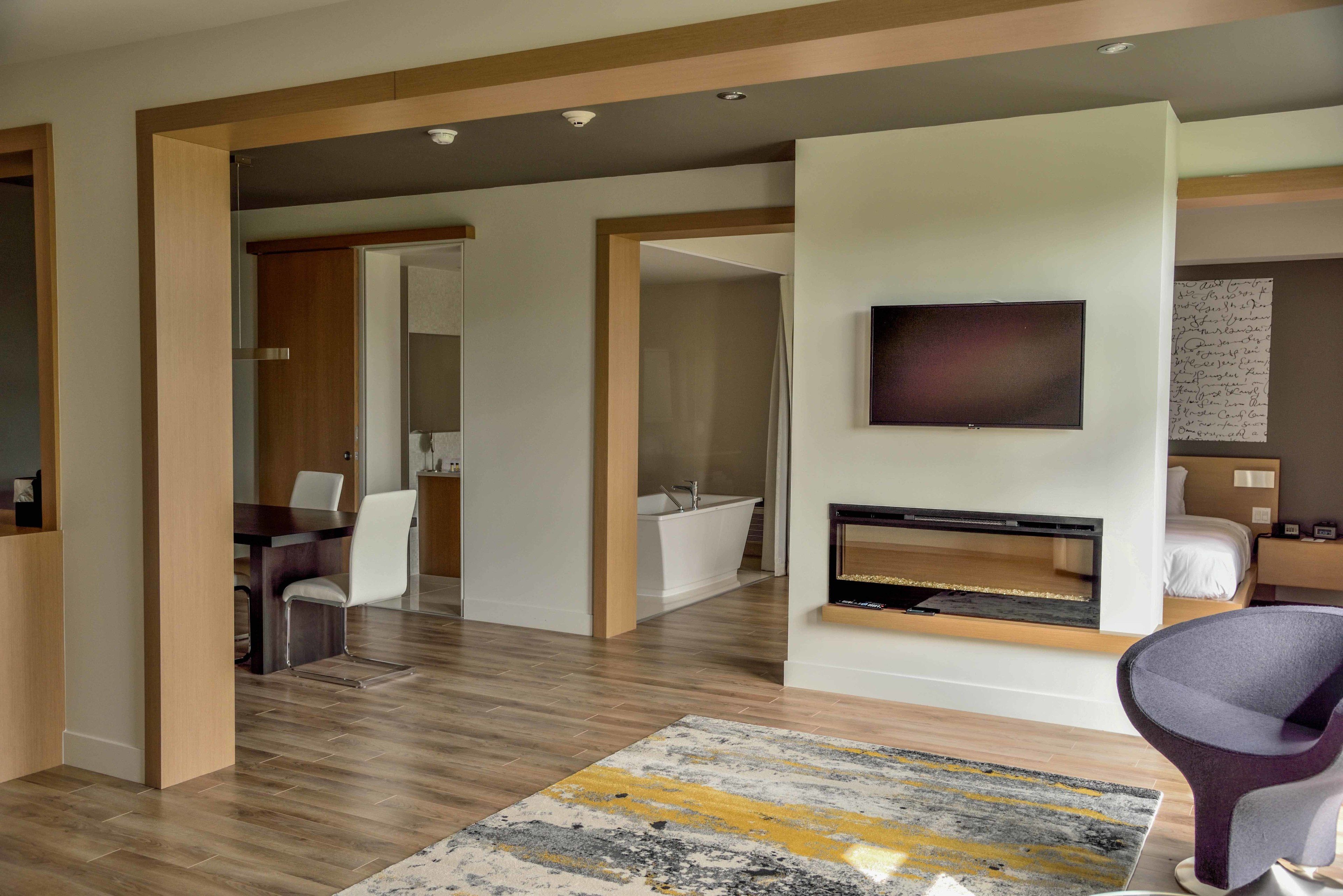 Ein modernes Hotelzimmer mit Holzboden, einem Flachbildfernseher über einem Kamin, einem Badezimmer mit Badewanne und einem Essbereich mit Tisch und Stühlen.