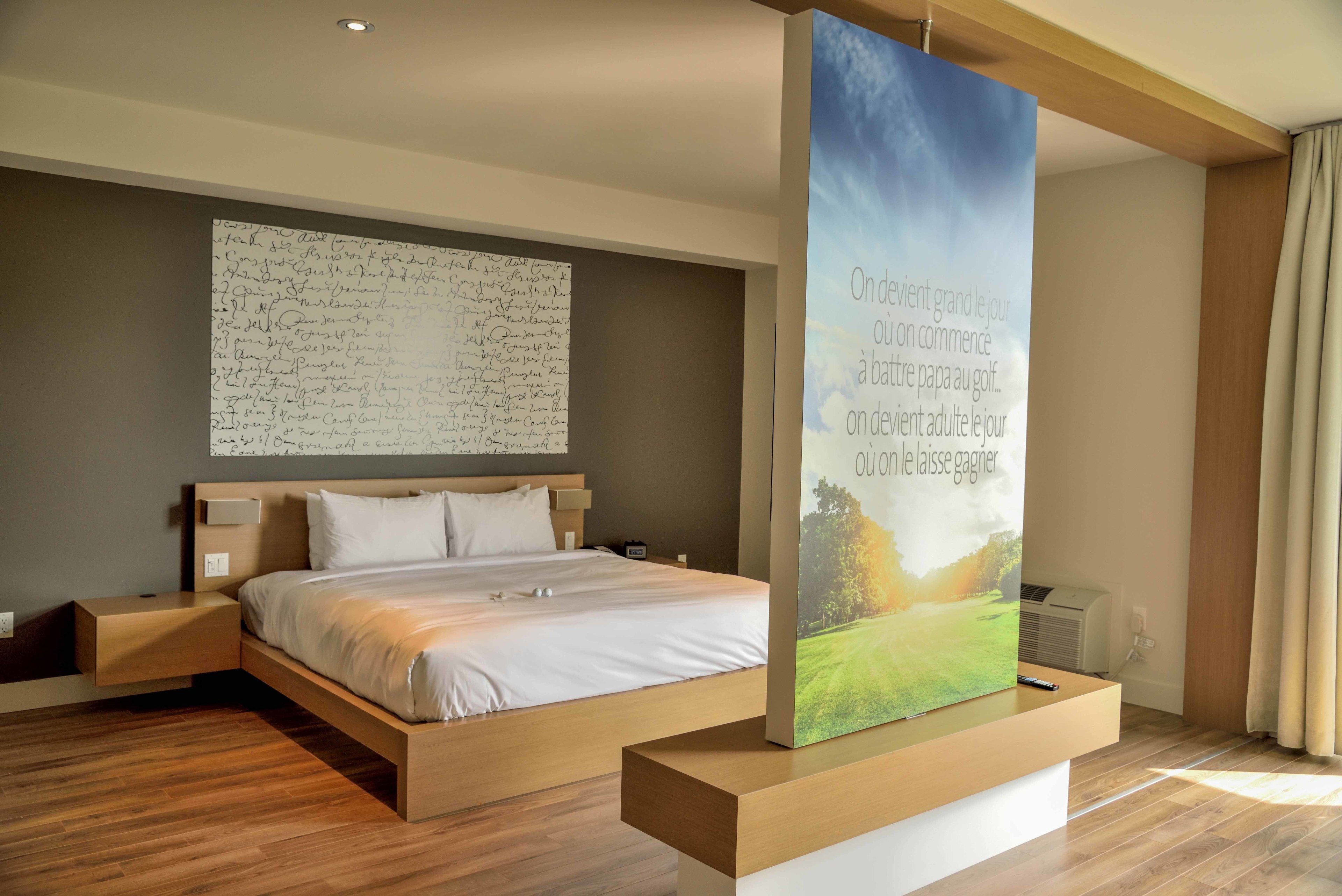 A modern hotel room with a large bed, a wooden nightstand, a wall-mounted air conditioner, and a large poster on the wall.