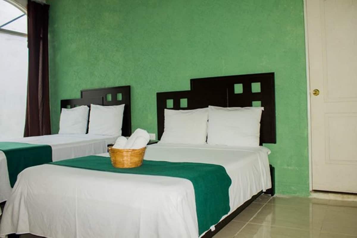 Une chambre avec deux lits, chacun recouvert d’une couverture verte et de draps blancs. On aperçoit un panier en osier sur le lit, au premier plan.