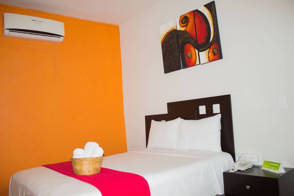 Une chambre d’hôtel avec un lit, un panier rempli de serviettes, une table de chevet avec un téléphone et un appareil de climatisation fixé au mur.