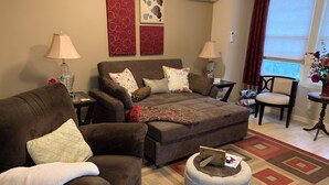 A cozy living room with a sofa, ottoman, and a small table with a magazine. There are two lamps, a vase with flowers, and artwork on the wall. - Kalmia Cottage (Junction City)