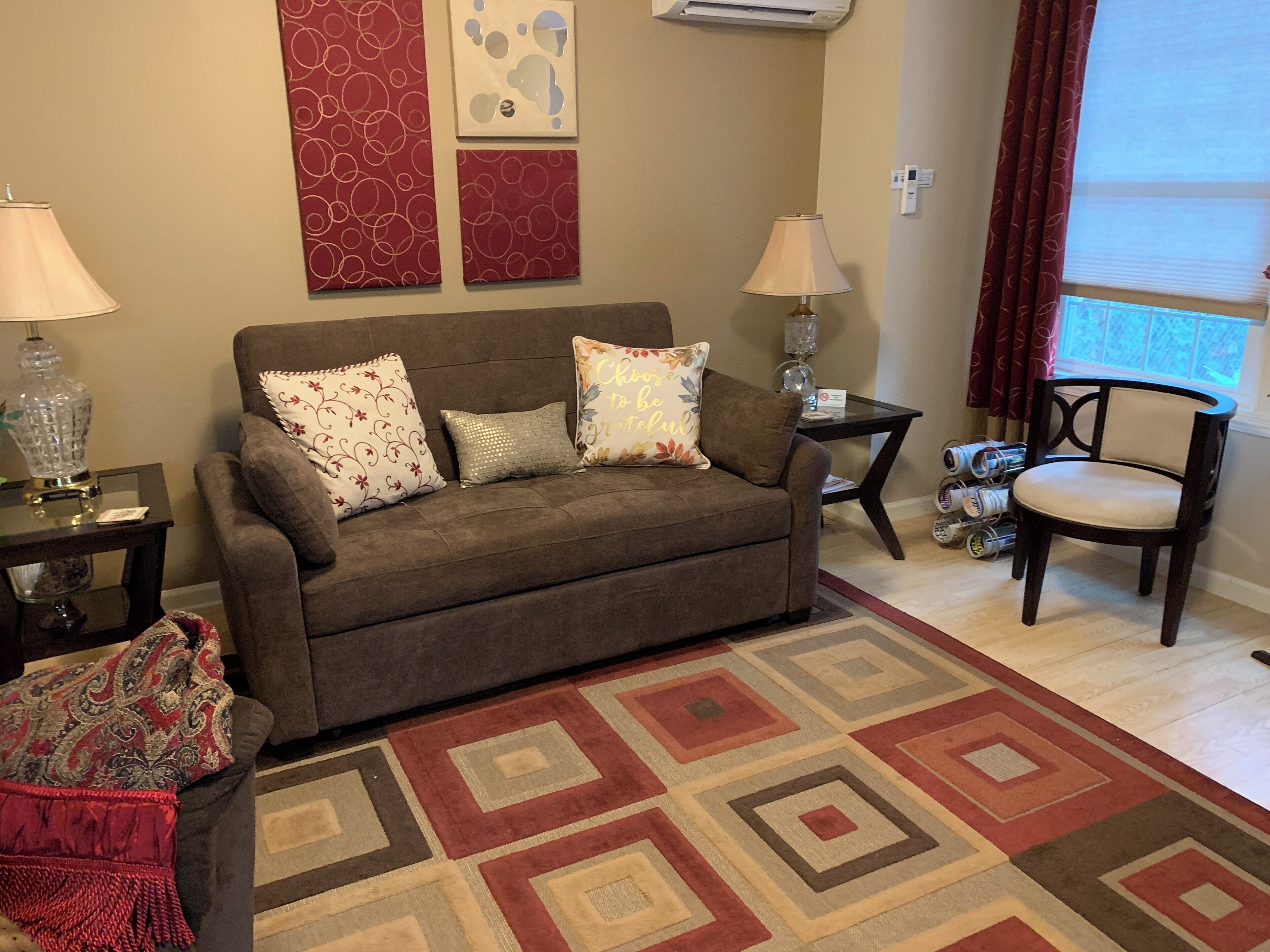 A living room with a brown sofa, patterned cushions, a decorative rug, a side table with a lamp, and a chair.