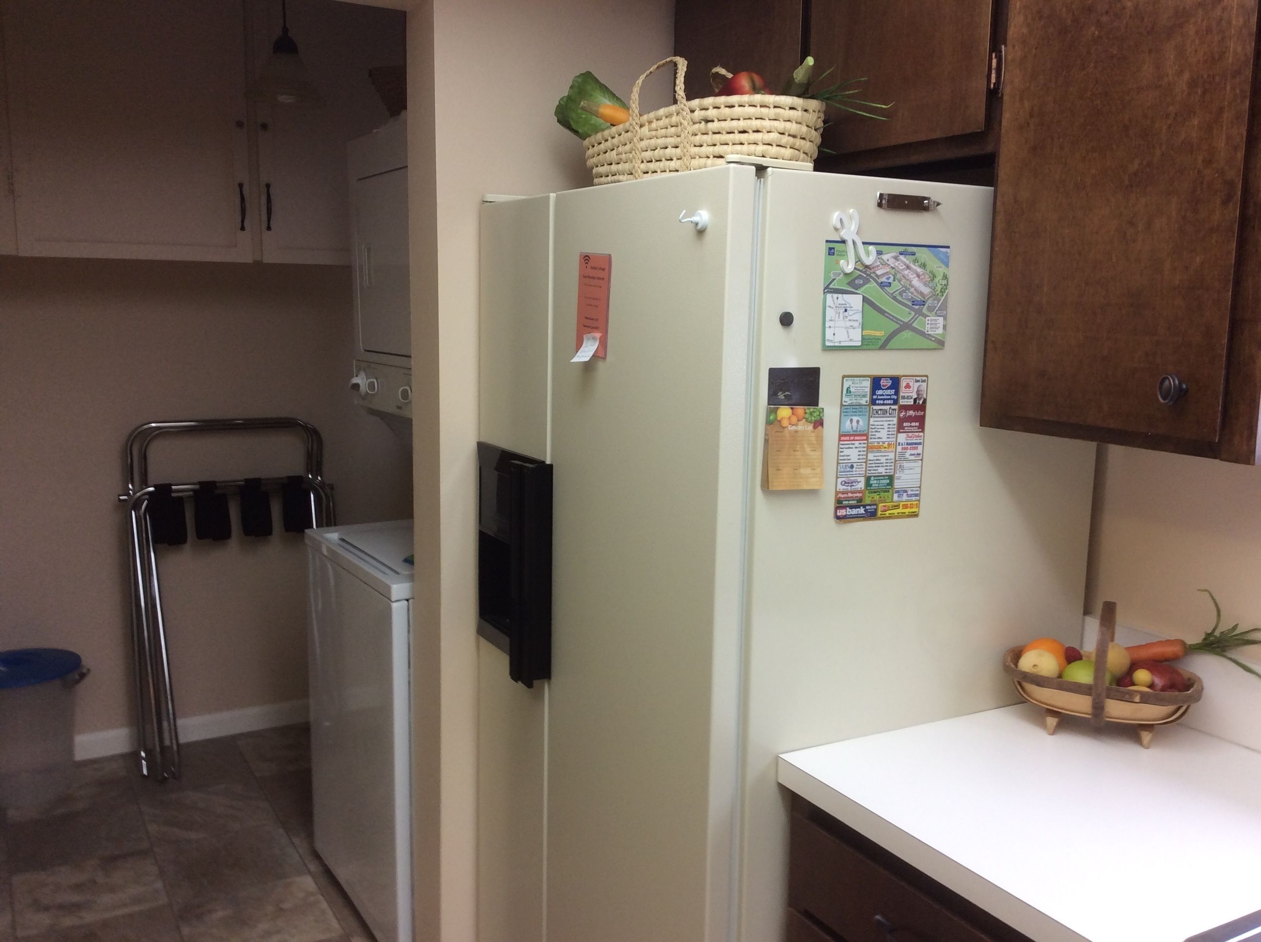 A kitchen with a refrigerator, a washing machine, and a small table with fruits.