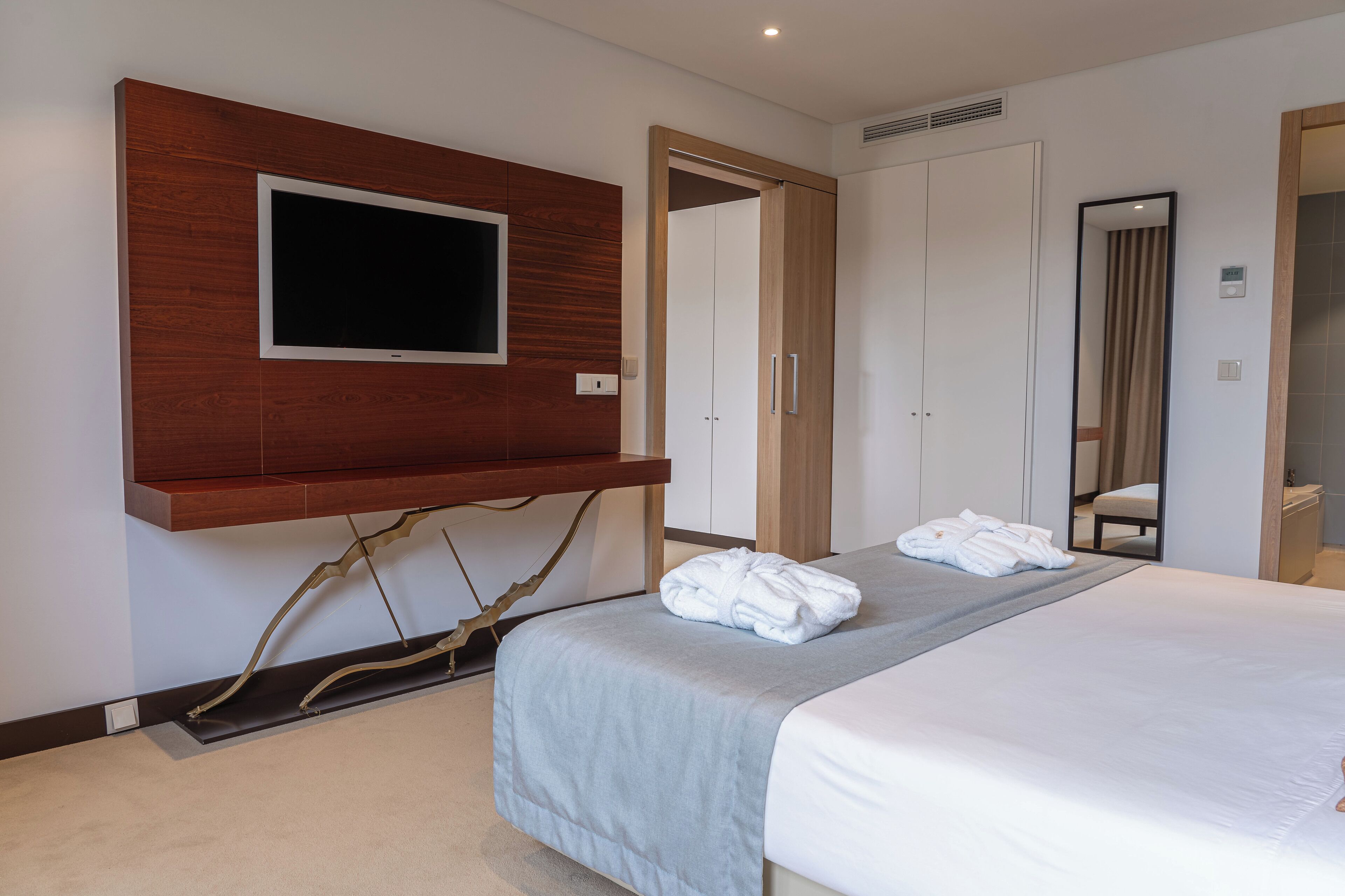A hotel room with a bed, a TV mounted on a wooden panel, a mirror, and a wardrobe.