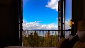 A room with a bed and a window overlooking a balcony with a view of trees and mountains.