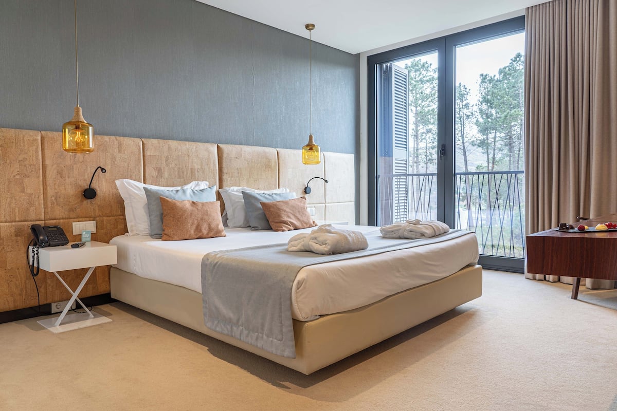 A modern hotel room with a large bed, bedside lamps, a small bedside table, and a view of the outdoors.