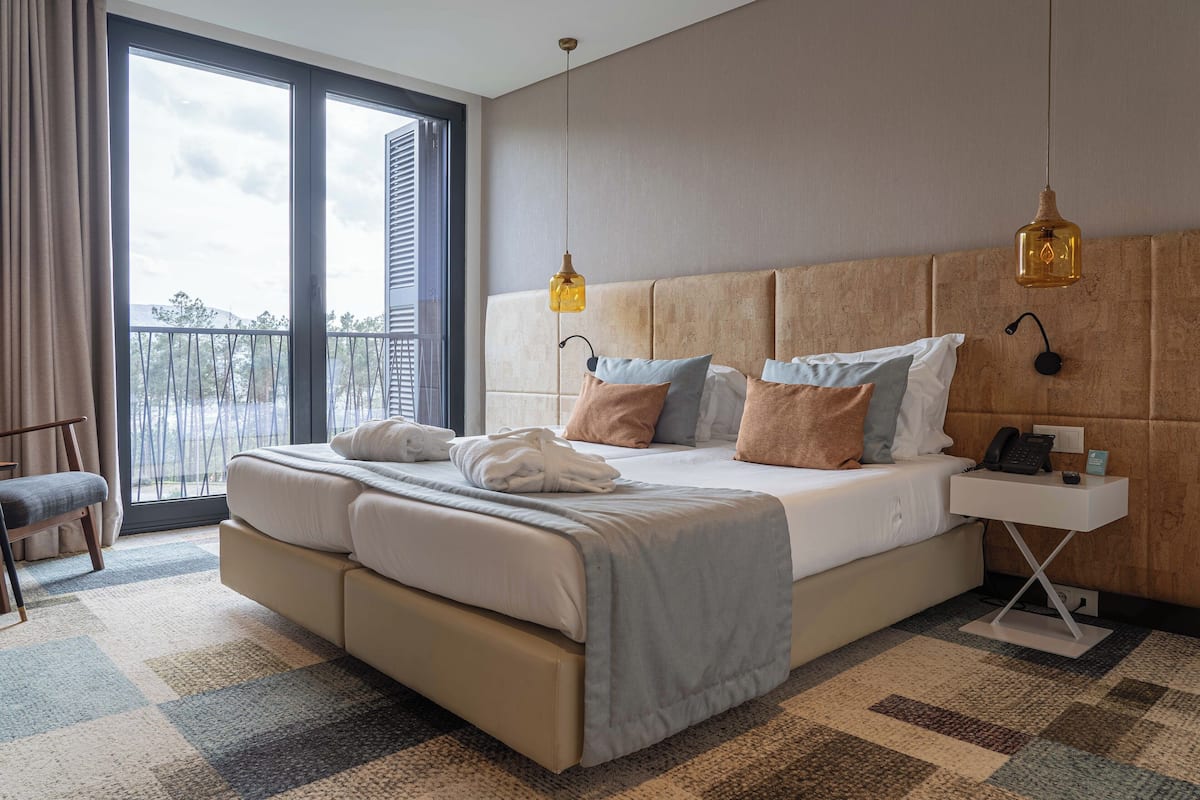 A modern hotel room with a large bed, two bedside tables, a view of the outdoors, and a balcony.