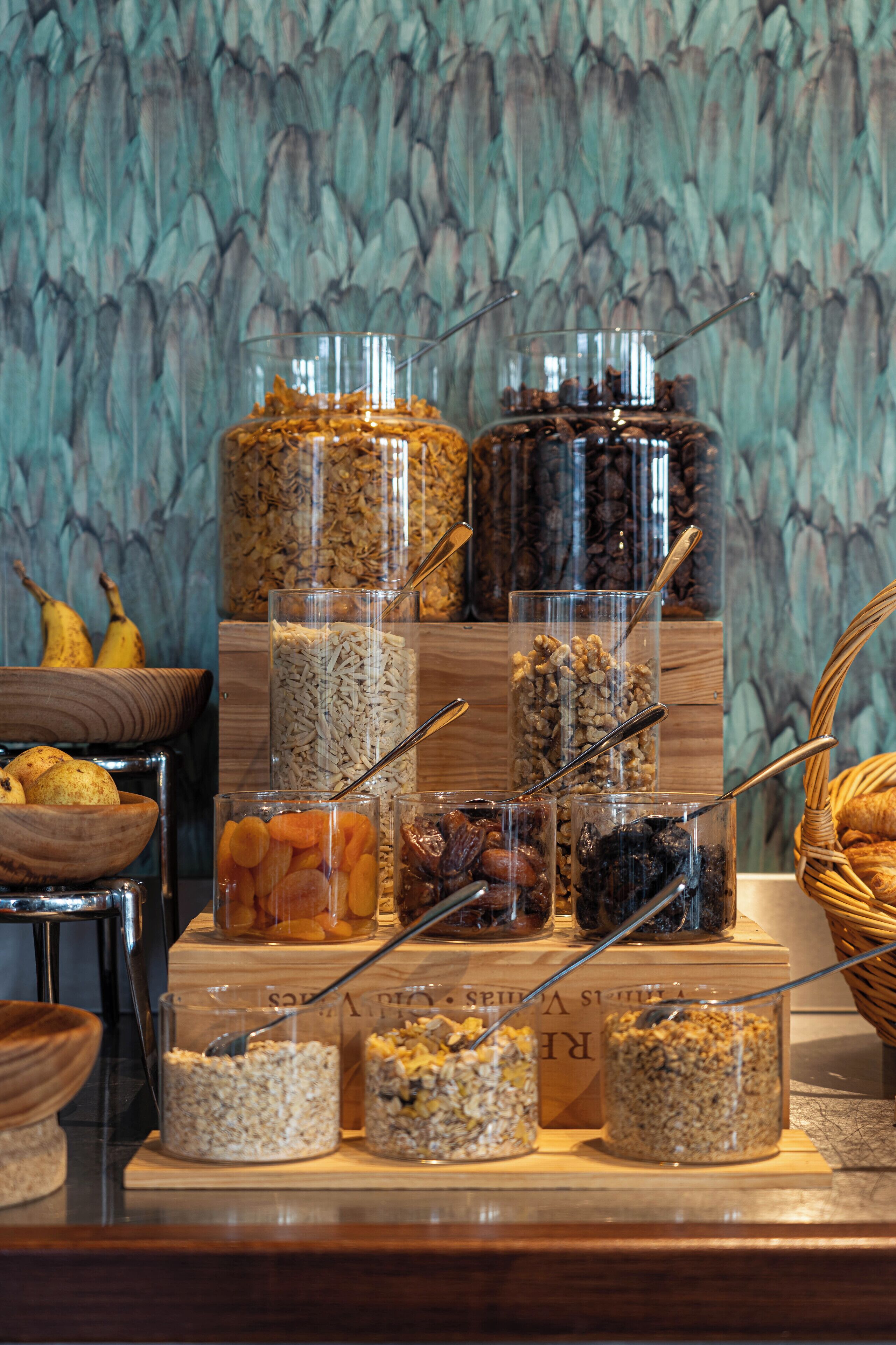 A variety of food items displayed in clear containers, including grains, nuts, and dried fruits, arranged on a wooden surface with a decorative background.