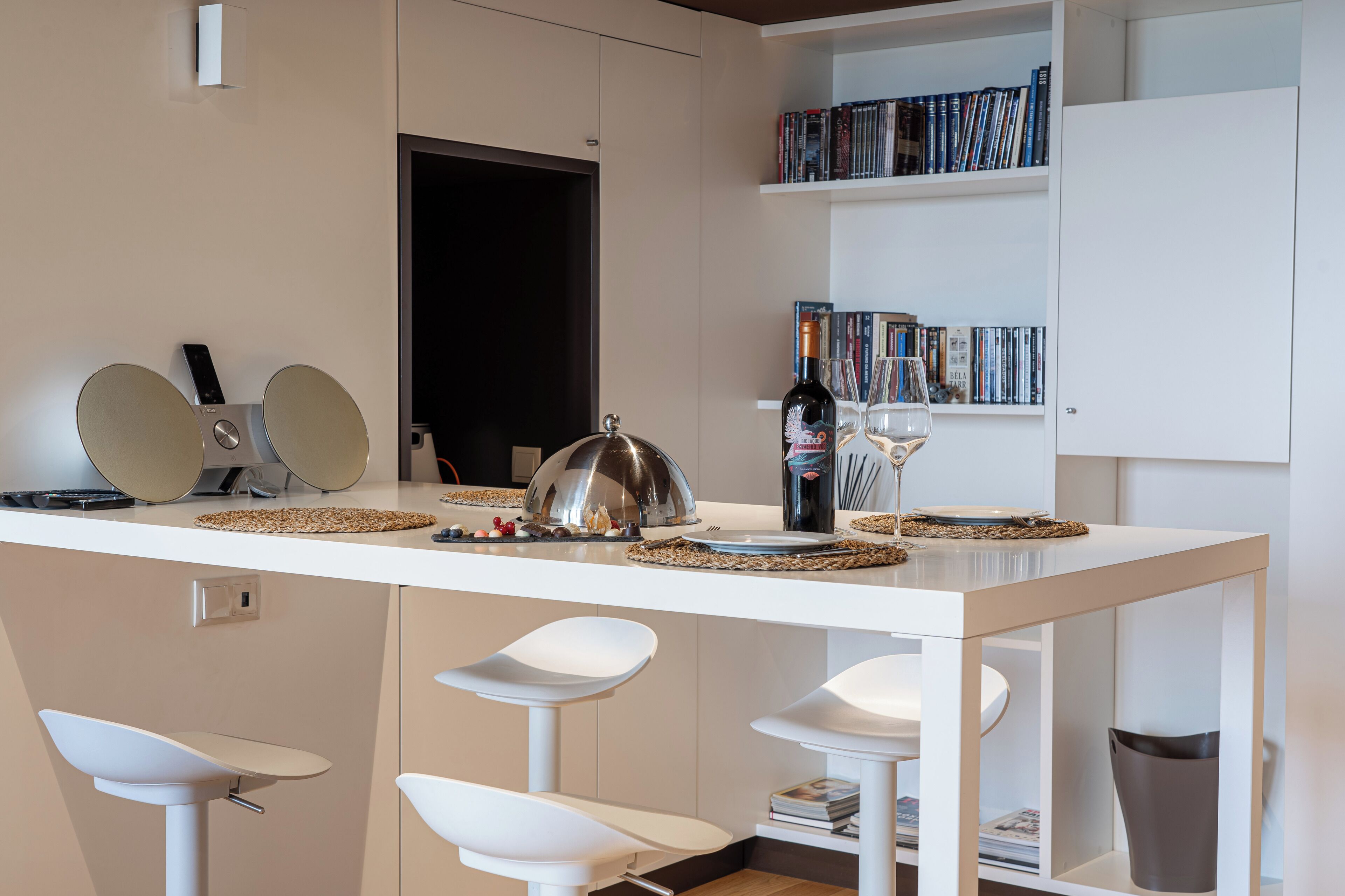 Una cocina moderna con encimera blanca, taburetes de bar, una botella de vino y una estantería llena de libros.