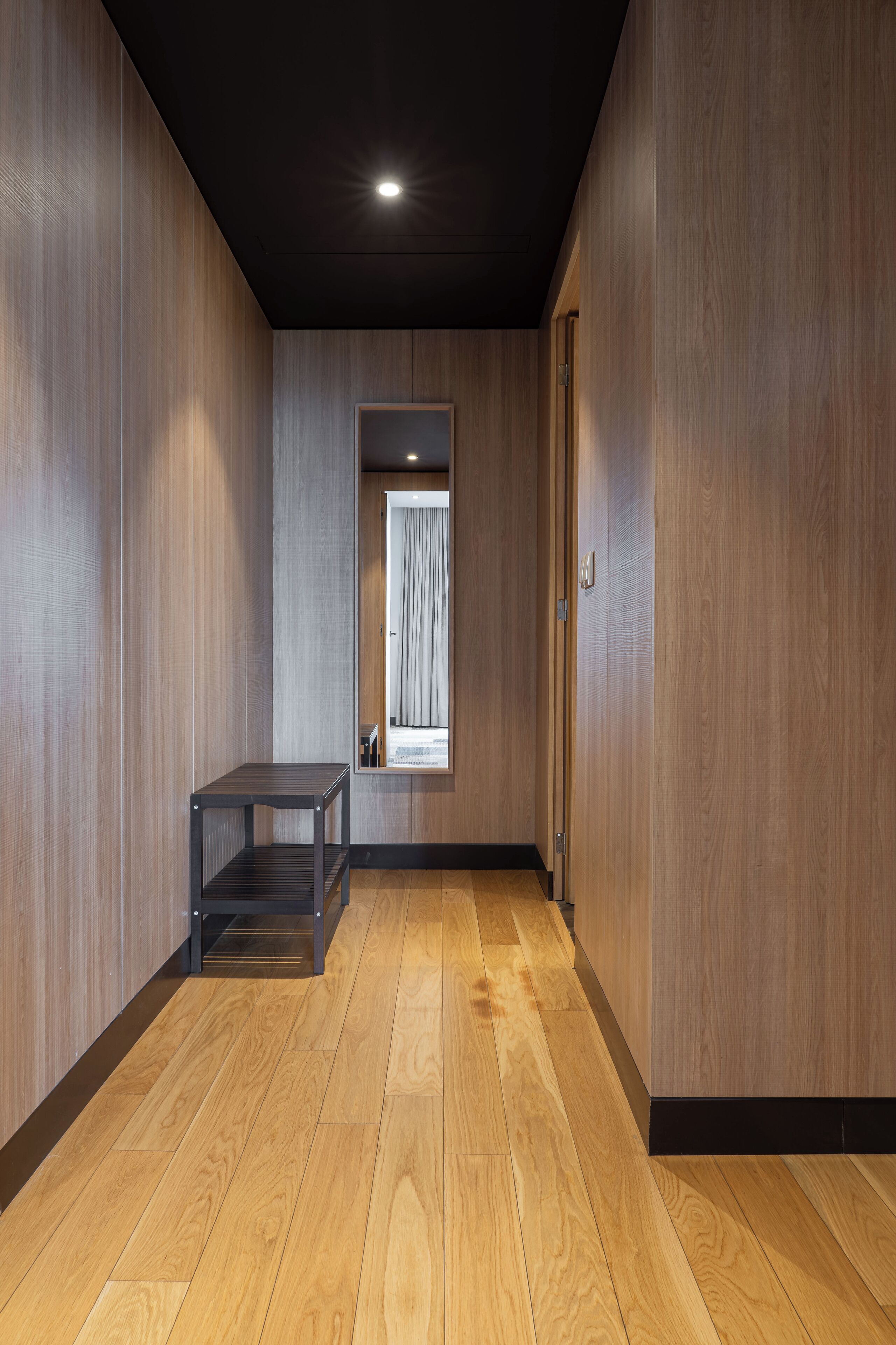 A hallway with wooden walls and a wooden floor, leading to a room with a bed and a window with curtains.