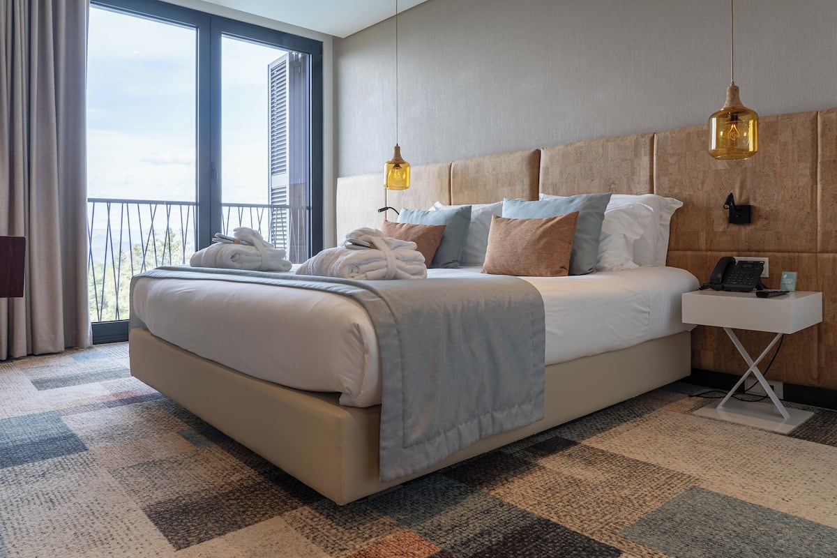 A hotel room with a large bed, a bedside table with a phone, and a view of the outdoors through a large window.