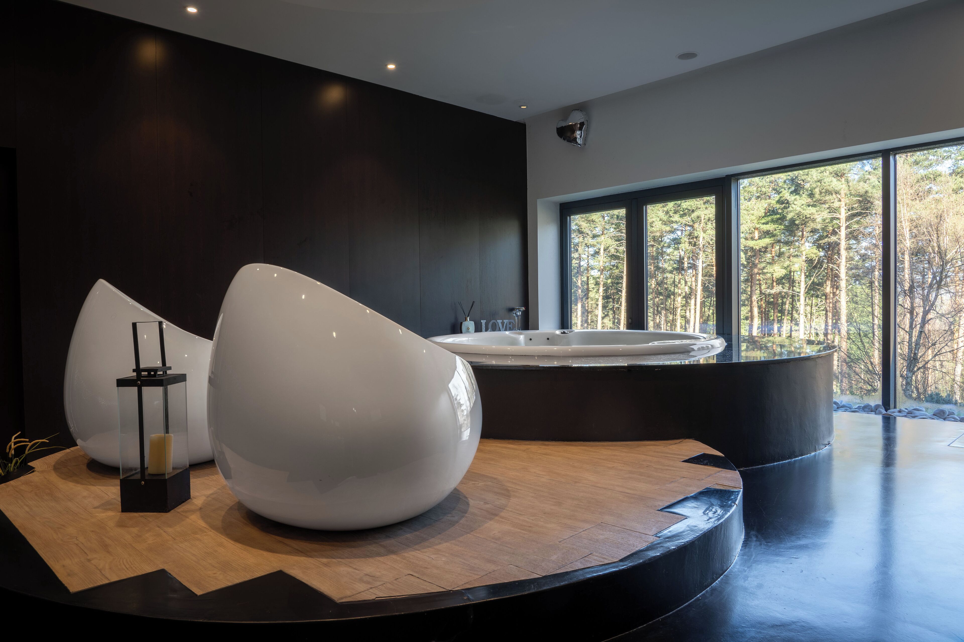A modern spa area with a large hot tub, a wooden floor, and a view of a forest through large windows.