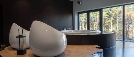 A modern spa area with a large hot tub, a wooden floor, and a view of a forest through large windows.